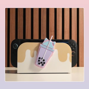 May include: A black and yellow games console with a cream-coloured stand featuring a dripping design. A purple and blue bubble tea charm is attached to the stand. The background is a wooden panel.