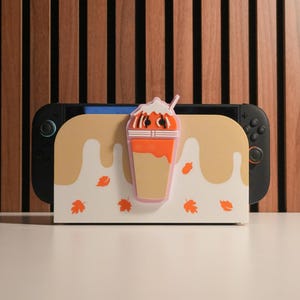 Puede incluir: Un soporte decorativo para consola de juegos con un diseño temático de otoño. El soporte tiene un fondo crema con un patrón de goteo y acentos de hojas naranjas. Una bebida de café de estilo caricatura está adjunta en la parte delantera.