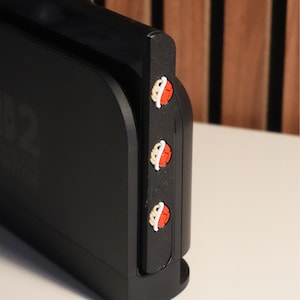 May include: A black gaming console with three decorative buttons featuring a red and white design. The console is positioned on a white surface, with a wooden-slat background visible.