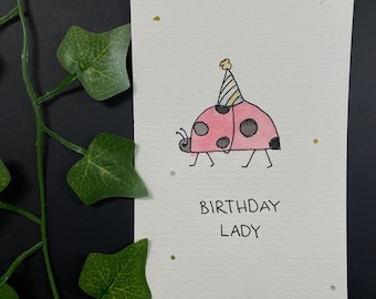 May include: A handmade birthday card featuring a pink ladybug with black spots and a party hat. The words "BIRTHDAY LADY" are written below the ladybug. The card is on a white background with gold and silver accents.