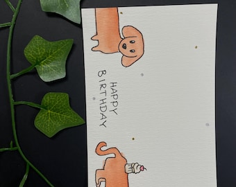 May include: A watercolor birthday card featuring two dachshunds. One dog is shown from the head up, and the other from the rear, holding a cupcake. The words "Happy Birthday" are written vertically. The card has gold and silver flecks.