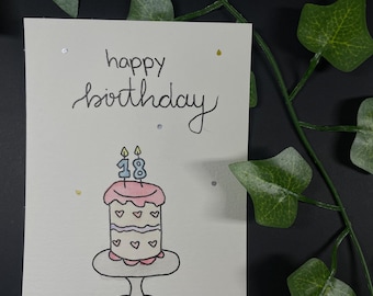 May include: Hand-drawn birthday card with the words "happy birthday" in cursive. The card features a cake with pink frosting, hearts, and candles that read "18". The card is decorated with gold and silver glitter.