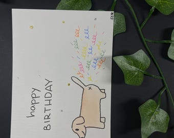 May include: A handmade birthday card with the words "happy BIRTHDAY" in black ink. A cartoon dog is drawn in the center, with colorful squiggles above it. The card is decorated with gold and silver dots.