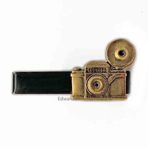 May include: A gold-toned tie clip featuring a vintage camera design with a black lens and a flash attachment. The camera is mounted on a black rectangular base.