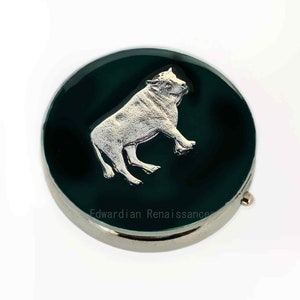 May include: A round, silver compact with a green enamel lid featuring a silver bull design. The compact has the text "Edwardian Renaissance" inscribed on the bottom.