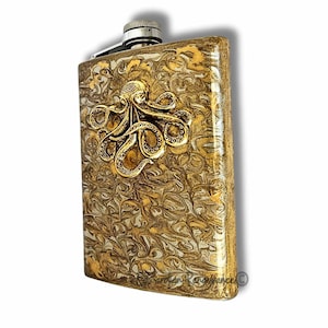 Puede incluir: Una petaca rectangular con un patrón de remolino dorado y blanco. Un emblema de pulpo dorado está centrado en la parte delantera. La petaca tiene una tapa plateada y un acabado de acero inoxidable. La petaca mide aproximadamente 10 cm de ancho y 13 cm de alto.