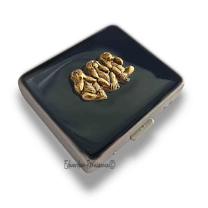May include: A black cigarette case with a silver trim and a gold three wise monkeys design. The monkeys are depicted covering their eyes, ears, and mouth.