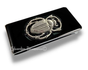May include: A black money clip with a silver scarab beetle design. The beetle is surrounded by a silver chain.