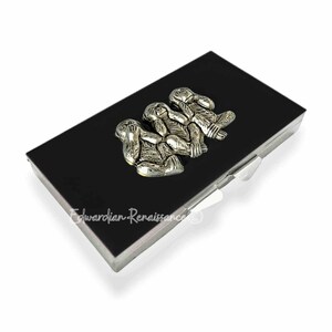 May include: A black rectangular box with a silver trim and a silver clasp. The top of the box features a silver sculpture of three monkeys in the "See no evil, hear no evil, speak no evil" pose.