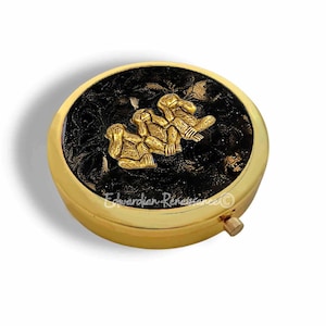 May include: A round, gold-colored pillbox with a black, textured top. The top features a gold-colored relief of three figures. The pillbox has a small, gold-colored clasp. The text "Edwardian Renaissance" is visible.