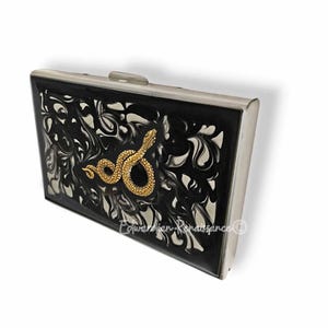 Op de afbeelding: Een rechthoekige zwarte clutch met een zilveren frame en sluiting. De voorkant heeft een wervelend zwart-wit patroon met een goudkleurige slangenversiering. Een stijlvol accessoire.