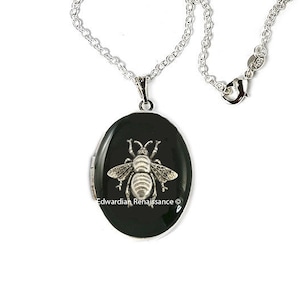 Puede incluir: Collar plateado con un colgante ovalado negro con un diseño detallado de abeja plateada. El colgante mide aproximadamente 3,8 cm de largo. La cadena tiene un cierre de mosquetón. Fondo blanco.