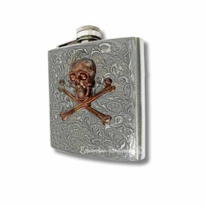 May include: A silver flask with a skull and crossbones design. The skull and crossbones are made of copper and are raised from the surface of the flask.