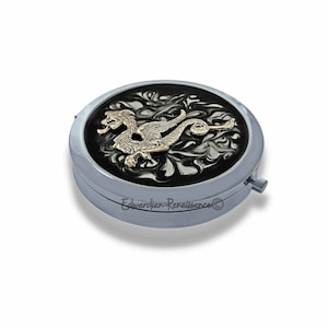 May include: A silver compact mirror with a black background and a gold dragon design. The mirror has a hinged lid and a small latch on the side.