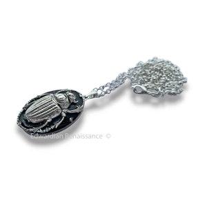 May include: A silver pendant necklace with a black oval charm featuring a detailed silver beetle design. The necklace is on a silver chain.