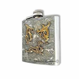 May include: A stainless steel hip flask with a silver and grey marbled pattern. Three gold-coloured dragon figures are attached to the front. The flask has a silver cap and hinge.