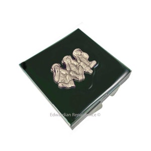 May include: A square, dark green compact mirror with a silver-colored relief of three monkeys covering their eyes, ears, and mouth. The compact has a silver-colored frame and a hinged opening. The words "Edwardian Renaissance" are printed on the bottom right.