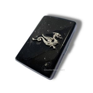 Könnte beinhalten: Eine schwarze rechteckige Hülle mit einem silbernen Drachenemblem. Die Hülle hat eine glänzende Oberfläche mit einem gesprenkelten Muster. Der Drache ist detailliert mit Flügeln und einem langen Schwanz.
