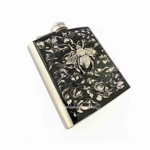 Puede incluir: Una petaca de acero inoxidable con un diseño de abeja plateada y un acabado marmoleado en blanco y negro.