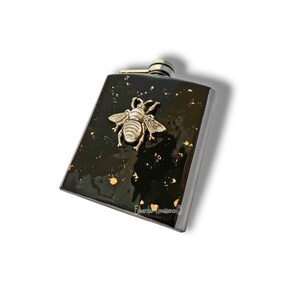 May include: Black stainless steel flask with a gold bee design. The flask has a black background with gold flecks.