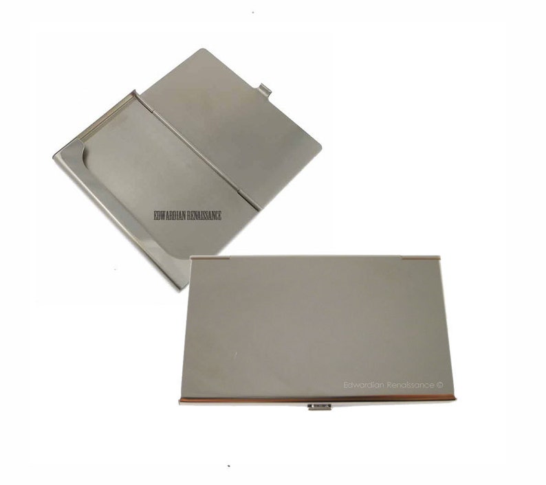 May include: A silver metal business card holder with a hinged lid. The holder is closed in the first image and open in the second image. The text "Edwardian Renaissance" is engraved on the holder.