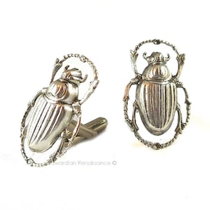Puede incluir: Un par de gemelos de plata con un diseño de escarabajo detallado. Los escarabajos están rodeados por un marco decorativo y ornamentado.