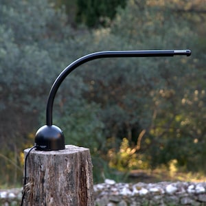 Puede incluir: Una lámpara de escritorio negra con un brazo curvo y una base redonda. La lámpara está colocada sobre un tocón de árbol. El fondo presenta follaje verde borroso y un muro de piedra. El brazo de la lámpara se extiende horizontalmente.