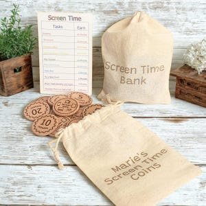 May include: A screen time reward system featuring a wooden chart with tasks and earned minutes, a collection of round wooden coins with engraved time values, and two beige drawstring canvas bags labeled Screen Time Bank and Marie's Screen Time Coins.
