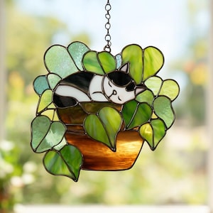 May include: Stained glass suncatcher featuring a sleeping black and white cat nestled in a pot of green leaves. The cat is curled up, appearing peaceful. The pot is brown, and the leaves are various shades of green. A chain is attached for hanging.