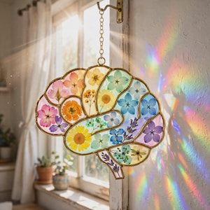 Può includere: Un acchiappasole a forma di cervello con fiori pressati di vari colori racchiusi in cornici di metallo dorato. L'oggetto è appeso a una catena e un gancio dorati, proiettando vivaci riflessi arcobaleno sulla parete adiacente.