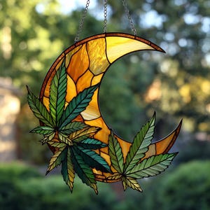 May include: Stained glass crescent moon with cannabis leaves. The moon is made of yellow and orange glass, with a brown border. Green and yellow cannabis leaves are attached to the moon. The piece is suspended by a chain.