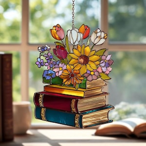 Pode incluir: Apanhador de sol em vitral com um buquê de flores coloridas em cima de uma pilha de livros. As flores incluem tulipas, girassóis e outras flores em tons vibrantes. Os livros são em tons de vermelho, amarelo e azul.