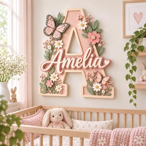 Könnte beinhalten: Ein großer, hellrosa Holzbuchstabe A als Wanddekoration mit dem Namen Amelia in weißer Schreibschrift. Der Buchstabe ist mit dreidimensionalen rosa und weißen Blumen, grünen Blättern und zwei Schmetterlingen in Rosatönen verziert.
