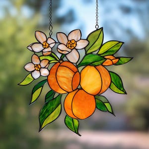 Può includere: Vetrata artistica raffigurante un grappolo di arance, fiori e foglie. Le arance sono di un arancione brillante, le foglie sono verdi e gialle, e i fiori sono bianchi con centri gialli. L'opera è sospesa da una catena.