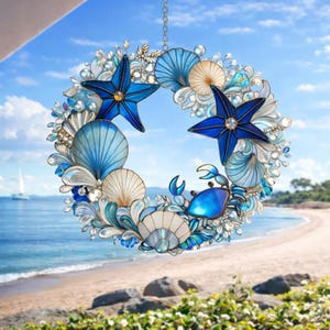 May include: A decorative wreath featuring a beach theme. The wreath is adorned with blue starfish, seashells, and a blue crab. The wreath is suspended by a chain and has a blue and white color scheme. The background shows a beach and ocean.