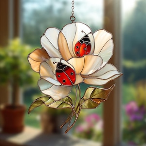 May include: Stained glass flower with two ladybugs. The flower has white and cream-colored petals with brown outlines. The ladybugs are red with black spots and black heads. The piece hangs from a chain and has green and gold leaves.