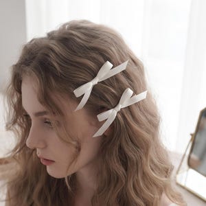 以下が含まれることがあります： オフホワイトのリボンヘアクリップ2つ。シンプルなリボンデザインで、滑らかなサテン調の生地が金属製のクリップに取り付けられています。長くウェーブのかかったライトブラウンの髪に留められ、ヘアスタイルにさりげない装飾的なアクセントを加えています。