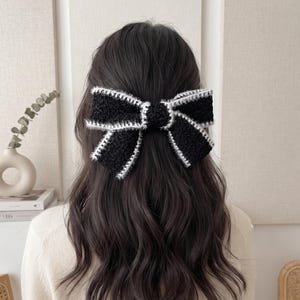 May include: A black hair bow with a textured, fuzzy surface and a white crocheted border. The bow is secured in long, dark, wavy hair. The design features a central knot with two wide, symmetrical loops extending outward on each side.