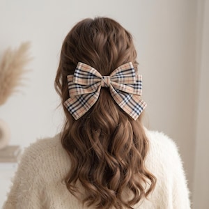 Könnte beinhalten: Eine große Haarschleife mit einem klassischen Karomuster in Beige-, Schwarz- und Rottönen. Die Schleife besteht aus mehreren Stofflagen, was ihr ein strukturiertes und voluminöses Aussehen verleiht. Sie ist an langem, welligem braunem Haar befestigt.