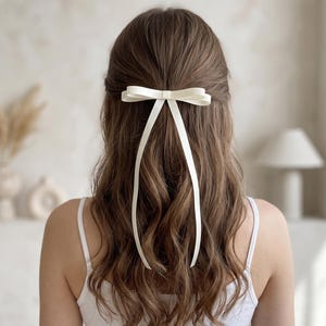 May include: A cream-coloured hair bow made of smooth ribbon, featuring a central knot and two long, trailing tails that hang down the back of the hair. The bow is secured horizontally, creating a clean, structured look with soft, flowing fabric.