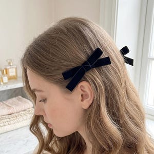 以下が含まれることがあります： 明るい茶色のウェーブヘアに付けられた2つの黒いベルベットのヘアリボン。各リボンは、2つのループと2つの垂れ下がった端を持つクラシックな結び目のデザインで、髪の毛をしっかりと固定するために頭の横に配置されています。
