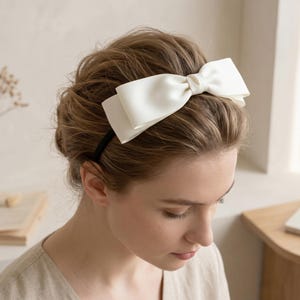 May include: A hair accessory featuring a large, smooth, off-white satin bow attached to a thin, black headband. The bow is positioned on the side of the head, showcasing a structured, multi-layered fabric design with a central knot detail.