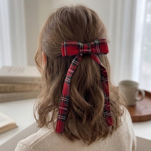 Könnte beinhalten: Eine rote Haarschleife mit langen Bändern. Die Schleife hat ein klassisches Tartanmuster in Rot, Schwarz, Grün und Weiß. Das Haaraccessoire wird im Haar getragen.
