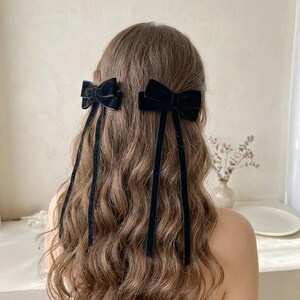 Può includere: Due fiocchi per capelli in velluto nero fissati a lunghi e sottili nastri di velluto. I fiocchi sono fissati su entrambi i lati della testa, con i nastri che pendono verticalmente lungo i capelli castani ondulati. Gli accessori hanno una consistenza liscia.