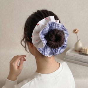 May include: A hair bun accessory with a dark brown bun, a white fabric layer with red cherry prints, and a blue and white gingham pattern. Designed to be worn in the hair.