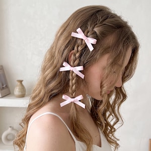 May include: Three small, light pink satin ribbon bows are attached to a single braided section of wavy, light brown hair. Each bow is tied in a classic style with two loops and two short tails, positioned at intervals along the length of the braid.