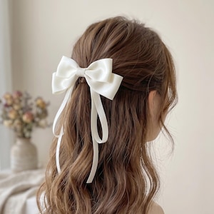 Puede incluir: Un lazo de satén blanco para el cabello con largas cintas sueltas. El lazo está centrado en el cabello, agregando un toque de elegancia. El cabello es ondulado y castaño, con el lazo como punto focal. Un accesorio para el cabello simple pero elegante.