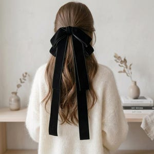 Puede incluir: Un accesorio para el cabello con lazo de terciopelo negro con largas cintas sueltas. El lazo está atado en la parte posterior de la cabeza, con las cintas cayendo sobre un cabello castaño largo y ondulado. La persona lleva un suéter blanco.