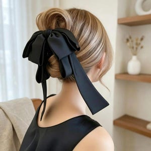 以下が含まれることがあります： 長いリボンの垂れ下がった大きな黒い布製リボンが付いたヘアクリップ。ブロンドのアップヘアに固定されており、マットな質感の黒いリボンが髪の色と鮮やかなコントラストを生み出しています。