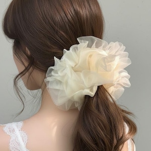 Peut inclure: Un chouchou crème en tulle froissé est porté en queue de cheval basse. L'accessoire pour cheveux est de couleur claire et neutre et ajoute une touche d'élégance à la coiffure. Les cheveux sont brun foncé.